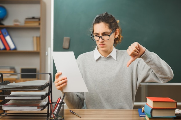 scontento che mostra i pollici verso il basso giovane insegnante maschio che indossa occhiali tenendo la carta seduto alla scrivania con gli strumenti della scuola in classe