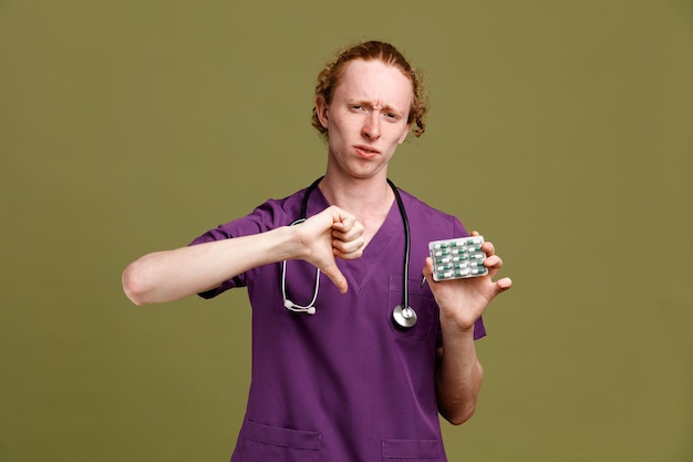 Scontento che mostra i pollici verso il basso giovane medico maschio che indossa l'uniforme con lo stetoscopio che tiene le pillole isolate su sfondo verde