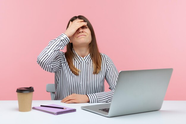 Sconvolto donna frustrata impiegato in camicia a righe che tocca il viso con la mano che fa gesto facepalm seduto al laptop difficoltà al lavoro errore Indoor studio girato isolato su sfondo rosa