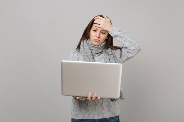 Sconvolto giovane donna in maglione grigio, sciarpa mettendo la mano sulla fronte, lavorando su computer pc portatile isolato su sfondo grigio muro. Stile di vita sano, consulenza sul trattamento online, concetto di stagione fredda.