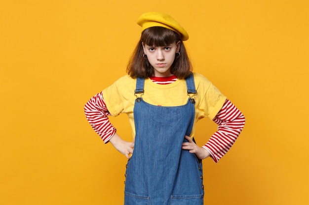 Sconvolto ragazza adolescente in berretto francese, prendisole in denim in piedi con le braccia sui fianchi isolato su sfondo giallo muro in studio. Persone sincere emozioni, concetto di stile di vita. Mock up spazio di copia.