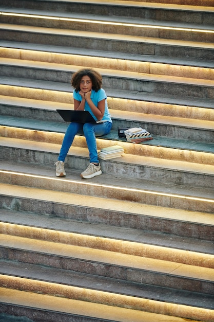 Scoolgirl adolescente riccia che si siede sui gradini all'aperto, leggendo il libro e scrivendo saggi sul laptop per lezioni di scrittura creativa