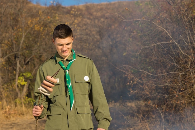 Scout che si prepara a cucinare salsicce su un fuoco in piedi guardando il fumo mentre si tiene le salsicce infilate su uno spiedino o un bastone