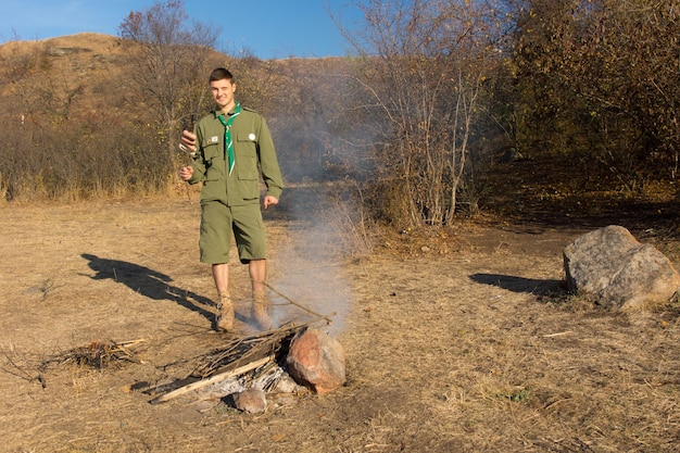 Scout o ranger che fa un fuoco di cottura in una radura nella vegetazione della prateria per cucinare il suo pranzo in piedi sorridendo alla telecamera