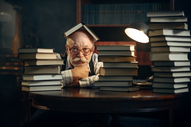 Scrittore anziano carino seduto al tavolo con una pila di libri in ufficio a casa. Il vecchio con gli occhiali scrive un romanzo di letteratura in una stanza con fumo, ispirazione