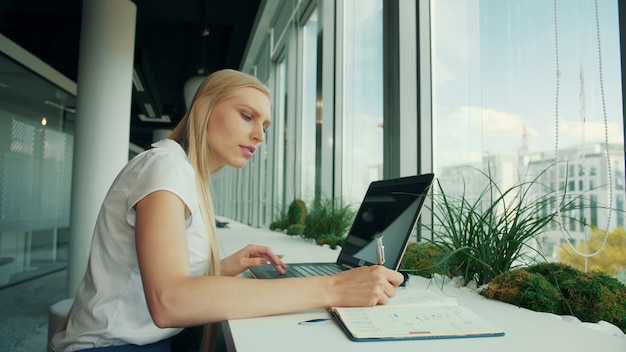 Scrittura della donna di affari sulla carta mentre sedendosi con il computer portatile alla tavola vicino alla finestra in ufficio moderno