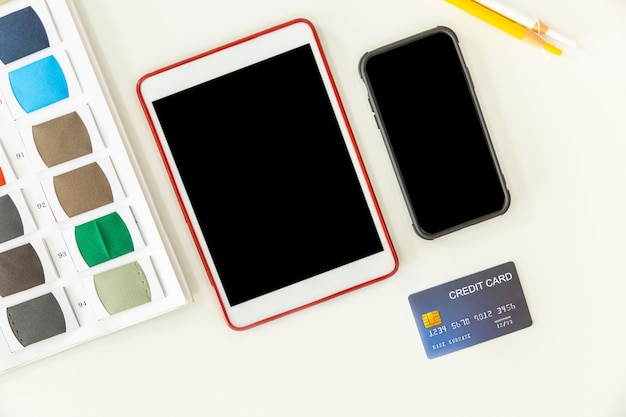 Scrivania bianca vista dall'alto con tablet e smartphone con schermo nero MOCKUP carta di credito e campione di stoffa