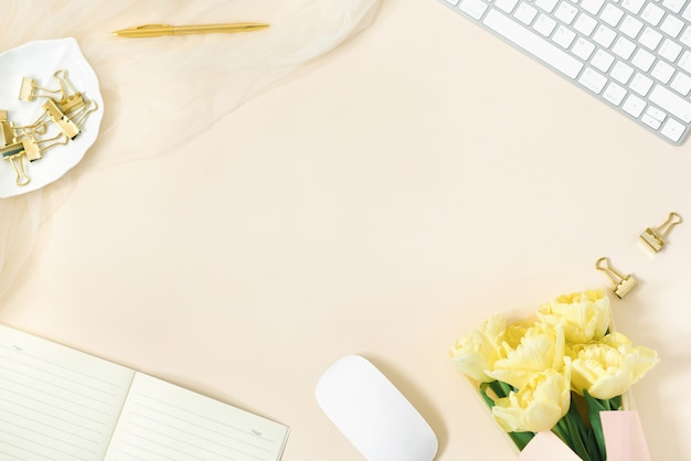 Scrivania da donna stilizzata, scrivania. Area di lavoro con un computer, un mazzo di tulipani gialli, appunti. Accessori moda femminile su uno sfondo beige chiaro. Vista dall'alto piatto