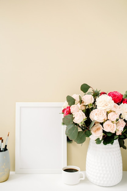 Scrivania da ufficio con cornice per foto, bellissime rose e bouquet di eucalipti davanti a uno sfondo beige pastello pallido