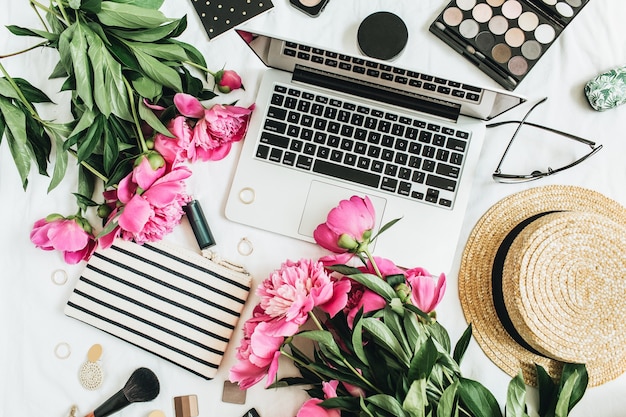 Scrivania da ufficio femminile alla moda piatta con laptop, fiori di peonia rosa, cosmetici, accessori. Fondo floreale di estate dell'area di lavoro di stile di vita di vista superiore.