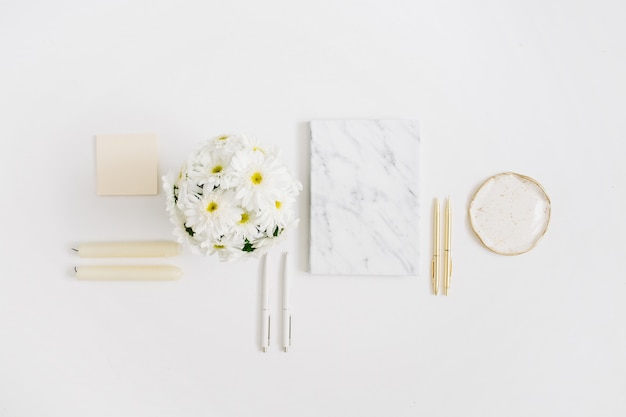 Scrivania da ufficio piatta. Area di lavoro della donna con bouquet di fiori di camomilla e diario in marmo su bianco