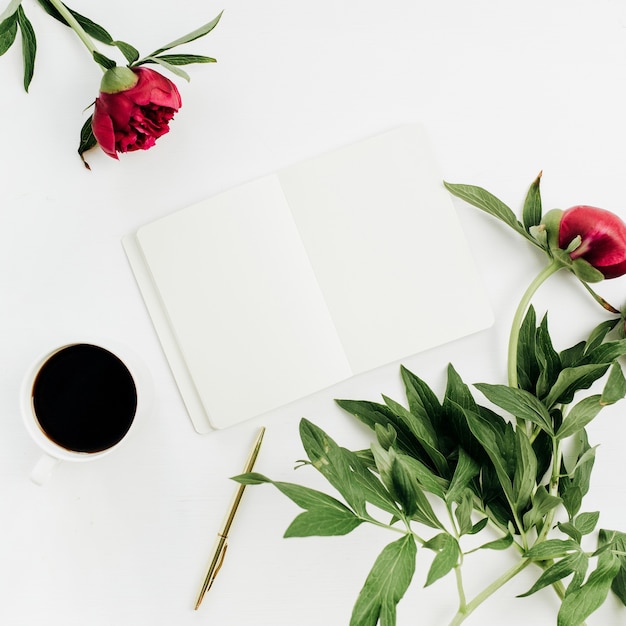 Scrivania minimale per l'home office con taccuino, caffè e fiori di peonia su sfondo bianco. Disposizione piatta, vista dall'alto