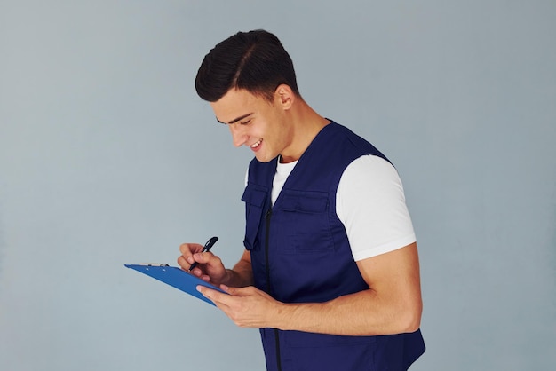 Scrive nel blocco note Lavoratore maschio in uniforme blu in piedi all'interno dello studio su sfondo bianco