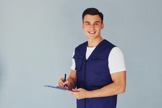 Scrive nel blocco note Lavoratore maschio in uniforme blu in piedi all'interno dello studio su sfondo bianco