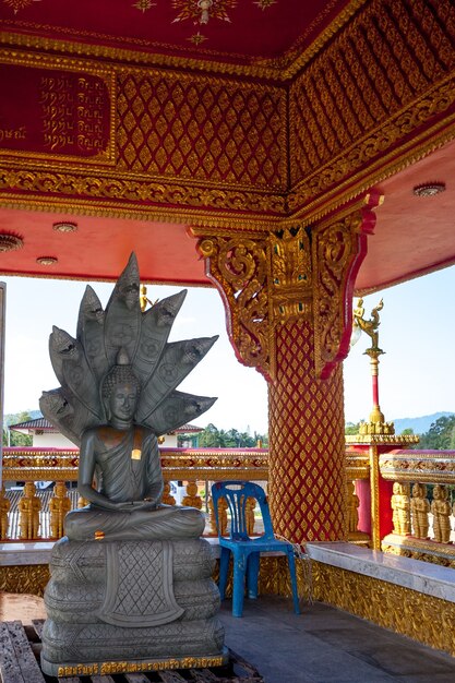 Scultura di Buddha nella posizione del loto sul trono con sette naga. La scultura è grigia sotto il tetto con un ornamento dorato. Testo dorato sotto.