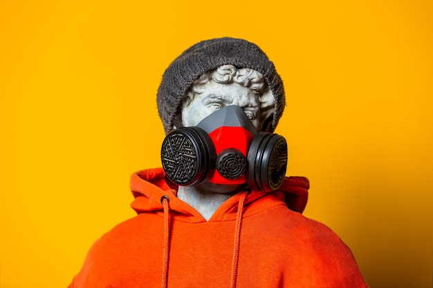 Scultura teenager in felpa con cappuccio arancione, maschera e cappello su sfondo giallo