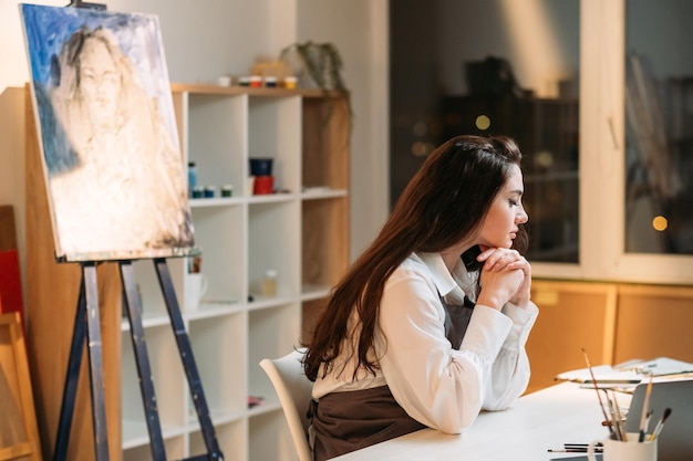 Scuola d'arte Pittore femminile Master class a casa Trova ispirazione Donna stanca e premurosa che tiene la testa sulle mani seduto a un tavolo con strumenti artistici in interni di uno studio luminoso