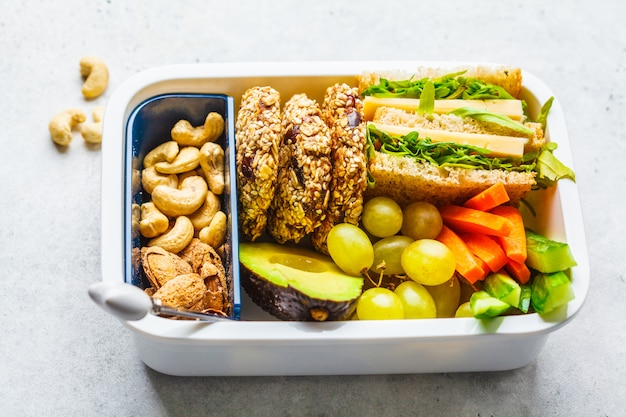 Scuola sana scatola di pranzo con sandwich, biscotti, frutta e avocado su sfondo bianco.