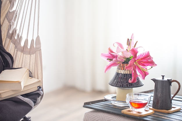 sedia pensile in stile boho con una pila di libri. Vassoio in legno con teiera e tazza di tè e bellissimi fiori di giglio.