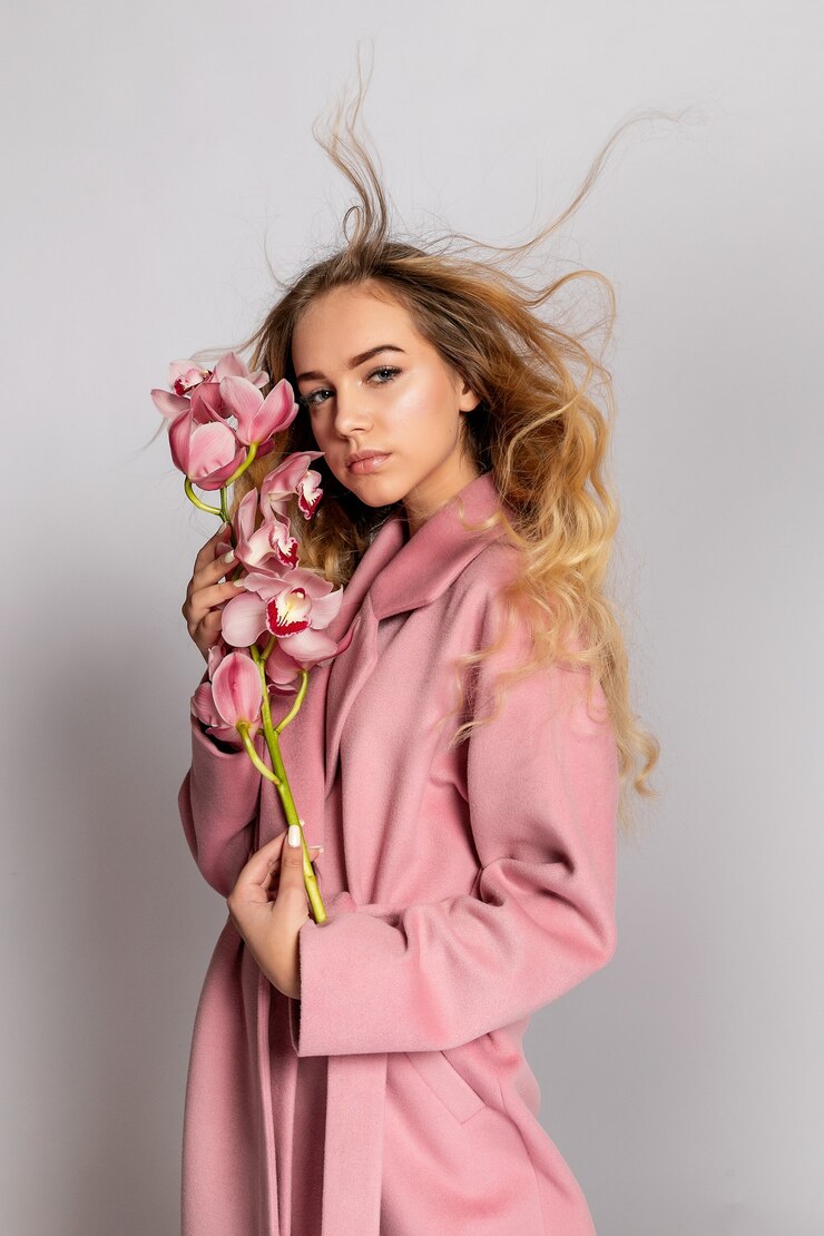 Foto seducente donna bionda in giacca rosa in posa in studio su sfondo grigio. ritratto di moda di un modello elegante in abito primaverile casual pastello. bella ragazza con pelle sana e orchidea ramificata