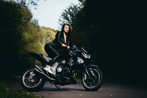 Seducente ragazza bruna con i capelli lunghi in una giacca di pelle nera si siede vicino a una moto moderna su uno sfondo di natura. Closeup ritratto di una donna sexy vicino a una costosa bici nera.