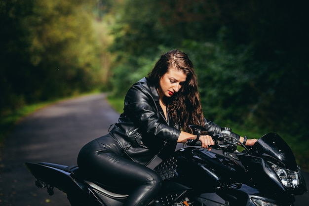 Seducente ragazza bruna con i capelli lunghi in una giacca di pelle nera si siede vicino a una moto moderna su uno sfondo di natura. Closeup ritratto di una donna sexy vicino a una costosa bici nera.