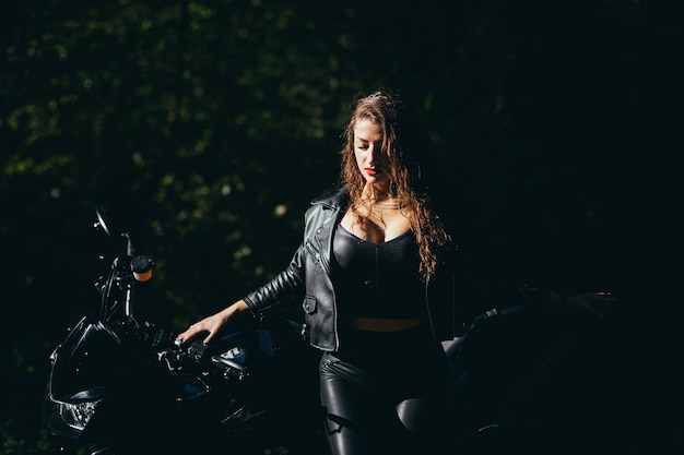 Seducente ragazza bruna con i capelli lunghi in una giacca di pelle nera si siede vicino a una moto moderna su uno sfondo di natura. Closeup ritratto di una donna sexy vicino a una costosa bici nera.