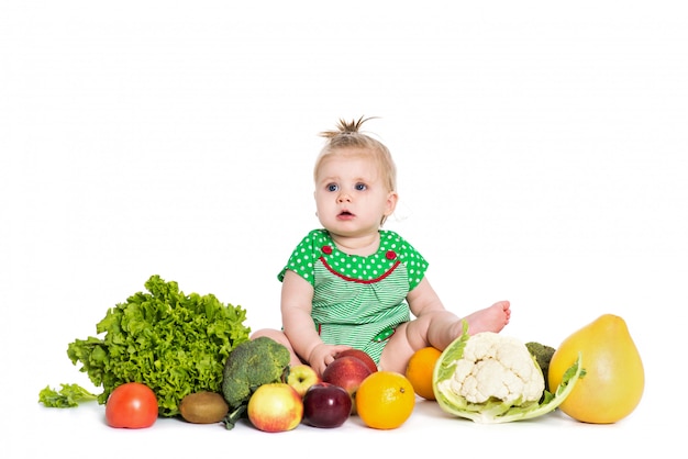 Seduta della neonata circondata dalla frutta e dalle verdure, isolata su bianco