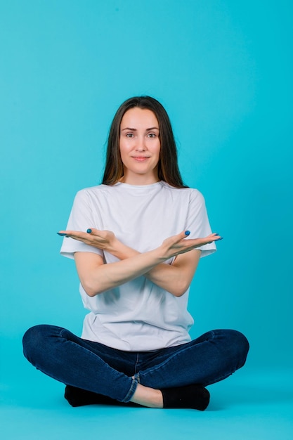 Seduta sul pavimento, la ragazza sta indicando con le mani incrociate su sfondo blu