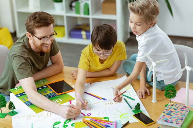 Seduto sul tavolo. Insegnante ridente positivo che lavora su poster colorati con i suoi studenti attivi