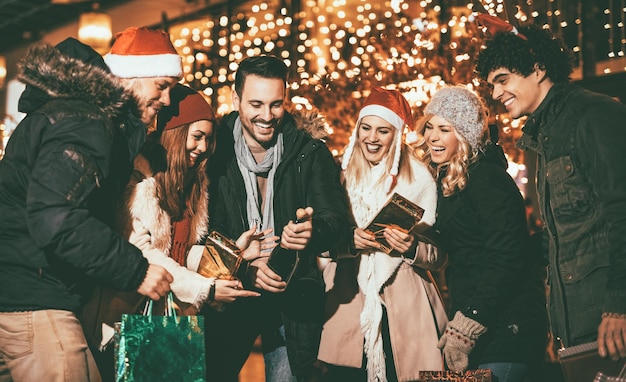 Sei giovani amici sorridenti in piedi fuori e con in mano molti sacchetti di carta della spesa, scatole e bottiglia di champagne nella notte della città.