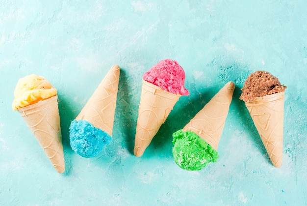 Selezione di vari brillanti gelati multicolori in coni gelato - cioccolato vaniglia mirtillo fragola pistacchio arancione, su luce blu soleggiata superficie, copia spazio vista dall'alto