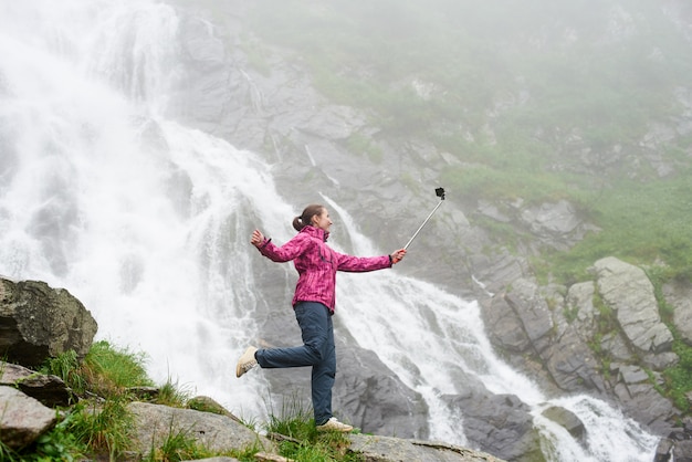 Selfie di fabbricazione femminile giovane che posa sulla roccia davanti alla cascata magnifica