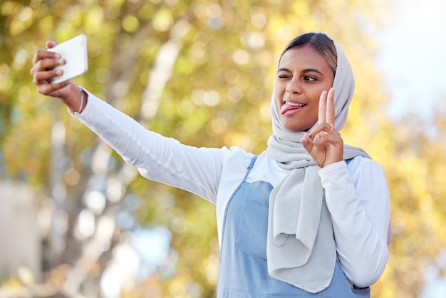 Selfie pace e donna islamica faccia sciocca all'aperto o fiduciosa con la felicità o la gioia dell'abbigliamento casual Signora musulmana in natura o smartphone per condividere foto social media o connessione con segno