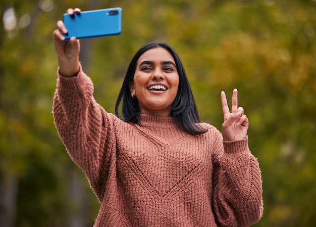 Selfie park e giovane donna pace e posa dello smartphone all'aperto nella natura e felice Gen Z dall'India sorriso e tecnologia divertimento e giovani che camminano fuori in giardino con gesto della mano