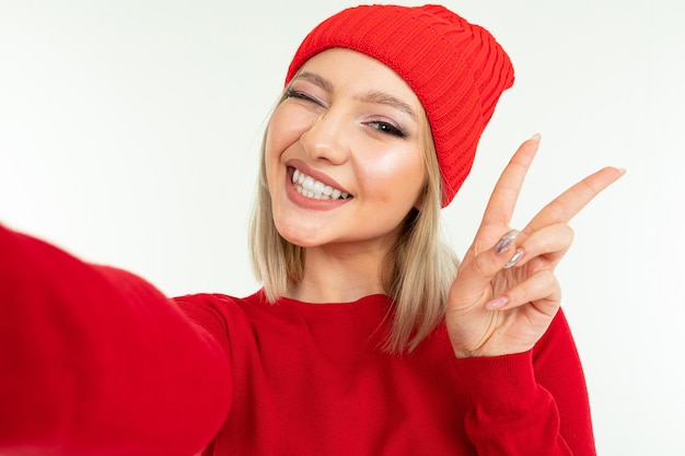 Selfie ragazza bionda con un cappello rosso e un maglione su uno sfondo bianco.