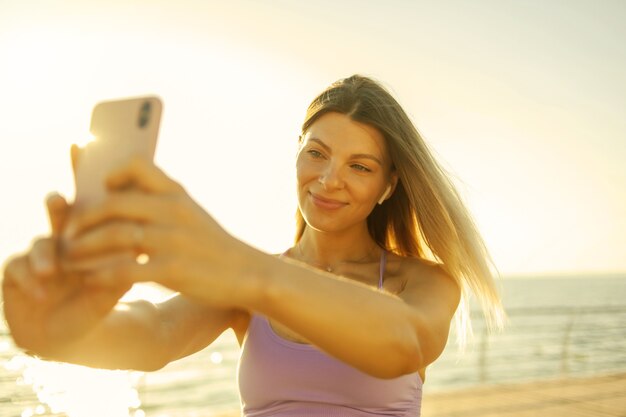 Selfie ritratto di una giovane donna bionda vestita con abiti sportivi sulla spiaggia all'alba