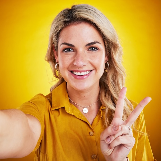 Selfie ritratto e pace con una donna su uno sfondo giallo in studio che sembra felice Sorriso del viso e gesto della mano con un attraente giovane modello femminile che mostra il simbolo v emoji o social media pov