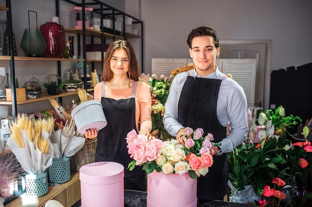 Sembrano belli e belli i giovani fioristi. Stanno al tavolo Giovane donna tenere piccola scatola grigia. Guy tocco fiori in scatola rosa.