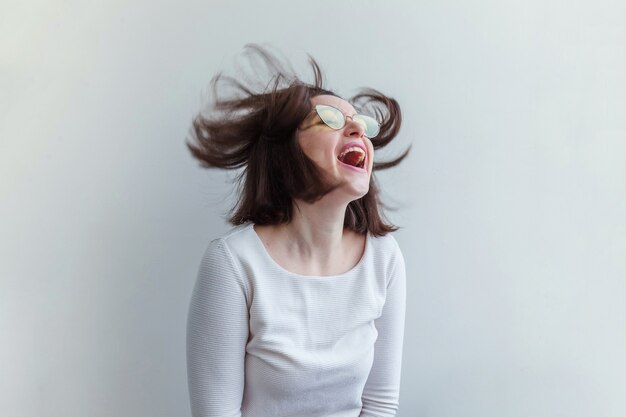 Semplice ritratto in studio di moda hipster sorridente ragazza bruna dai capelli corti in occhiali da sole gialli alla moda isolati su priorità bassa bianca. Divertente giovane donna intelligente positiva con gli occhiali che si diverte a sorridere