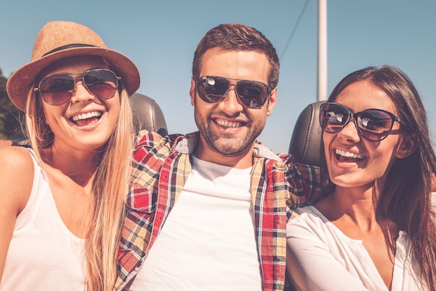 Sempre pronto a divertirti. Tre giovani felici che guardano la macchina fotografica e sorridono mentre si godono il viaggio in decappottabile