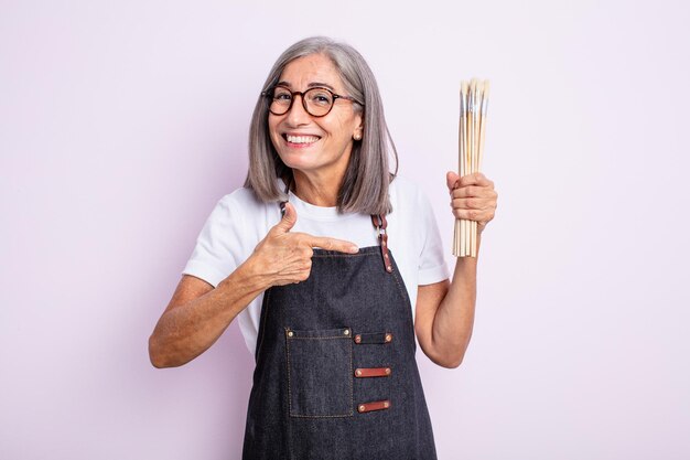 Senior bella donna che sembra eccitata e sorpresa indicando il lato. artista con i pennelli