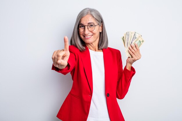 Senior bella donna sorridente e dall'aspetto amichevole, mostrando il numero uno. concetto di affari e banconote