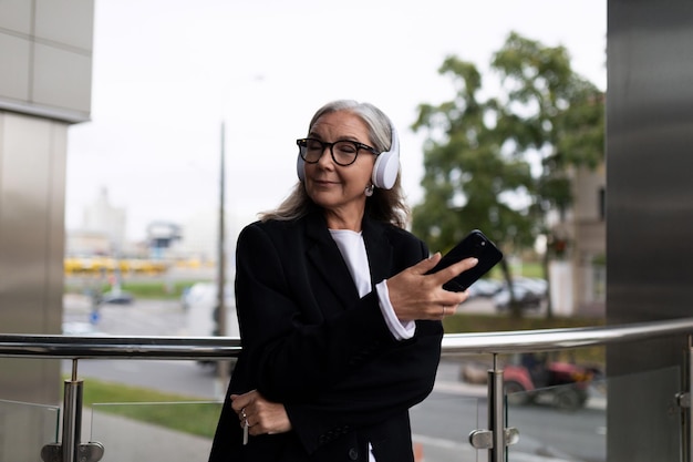 Senior donna alla moda che ascolta con piacere la musica in cuffia con il telefono cellulare nelle mani all'esterno