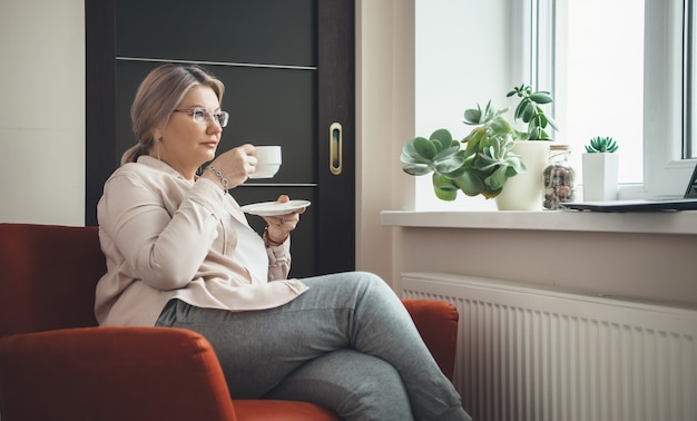 Senior donna caucasica con gli occhiali seduto in poltrona e bere un caffè mentre si indossano occhiali e si guarda una lezione sul computer portatile