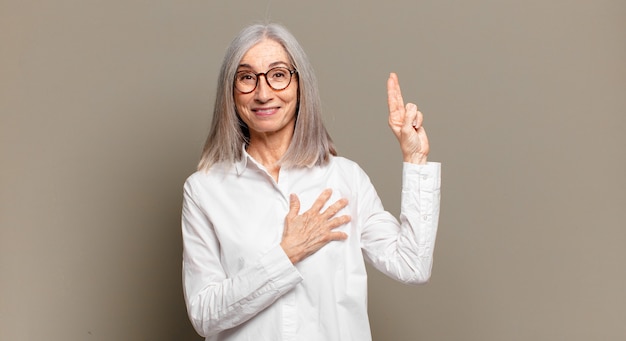 Senior donna che sembra felice, fiduciosa e degna di fiducia, sorridente e mostrando segno di vittoria, con un atteggiamento positivo