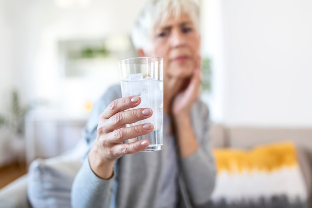 Senior donna con denti sensibili e mano che tiene un bicchiere di acqua fredda con ghiaccio. concetto di assistenza sanitaria. Donna matura che beve una bevanda fredda, un bicchiere pieno di cubetti di ghiaccio e sente mal di denti, dolore