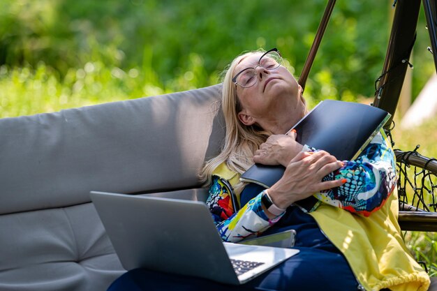 Senior donna con laptop stanco rilassante in giardino sul divano a dondolo tempo libero e concetto di vacanza