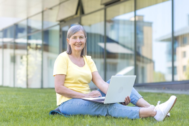 Senior donna dai capelli grigi felice seduta sull'erba e guardando la telecamera che lavora su un laptop fuori dall'ufficio