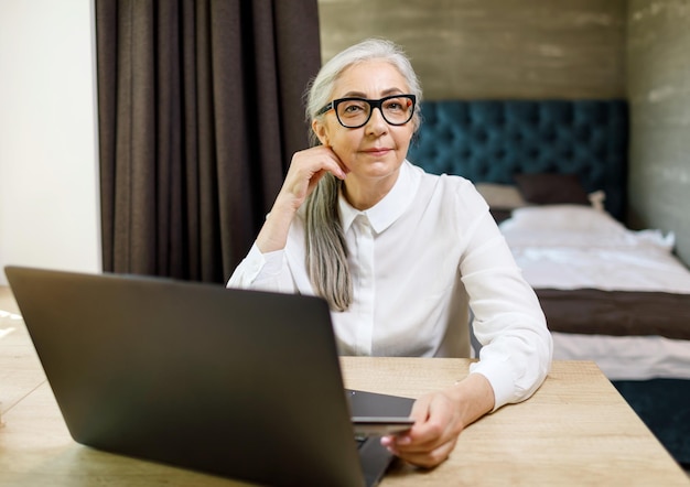 Senior donna dai capelli lunghi che indossa occhiali seduto con il computer portatile al tavolo nel suo appartamento confortevole